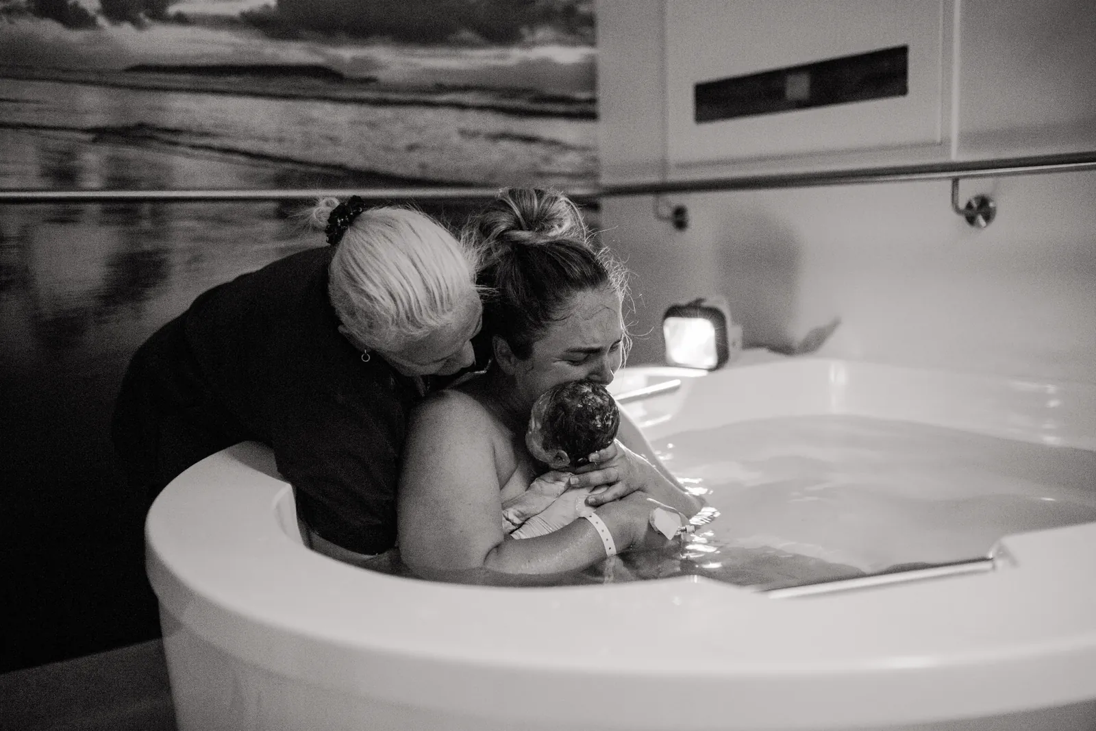 A mother holds her newborn in an AQVA maternity bath as her support person embraces her from behind