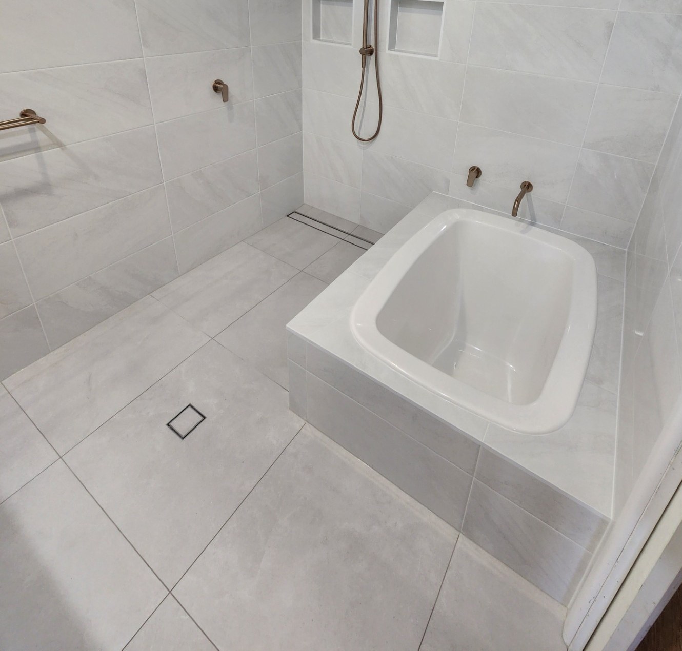 A deep white insert bath set into a tiled hob in a fully tiled wet room, with wall-mounted tapware and a hand shower