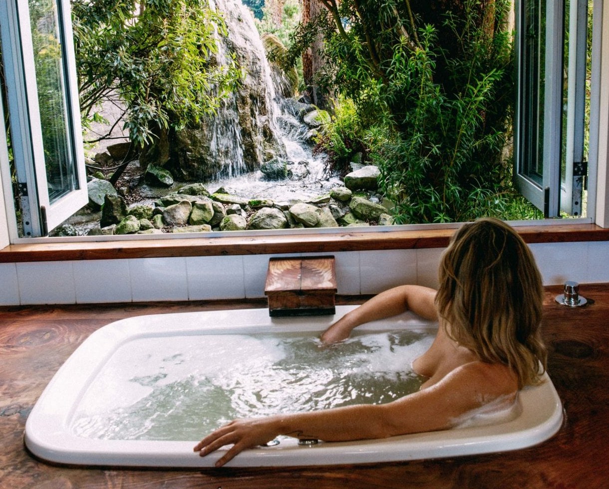A person leaning back in an insert spa with the jets running, set in a timber surround below an open window onto a garden and waterfall